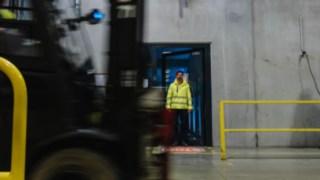Ohutusriietes mees astub uksest välja tehasehalli. Tõstuk sõidab mööda. Ohutusriietes mees astub uksest välja tehasehalli. Tõstuk sõidab mööda.